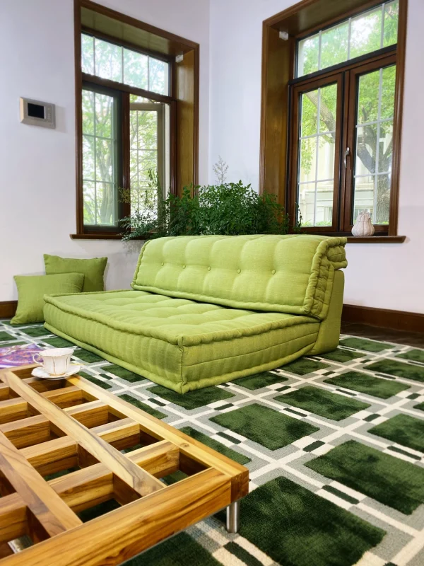 Close-up of a French green daybed with tufted cushions on a modern patterned rug, accompanied by a wooden coffee table and cup of tea.