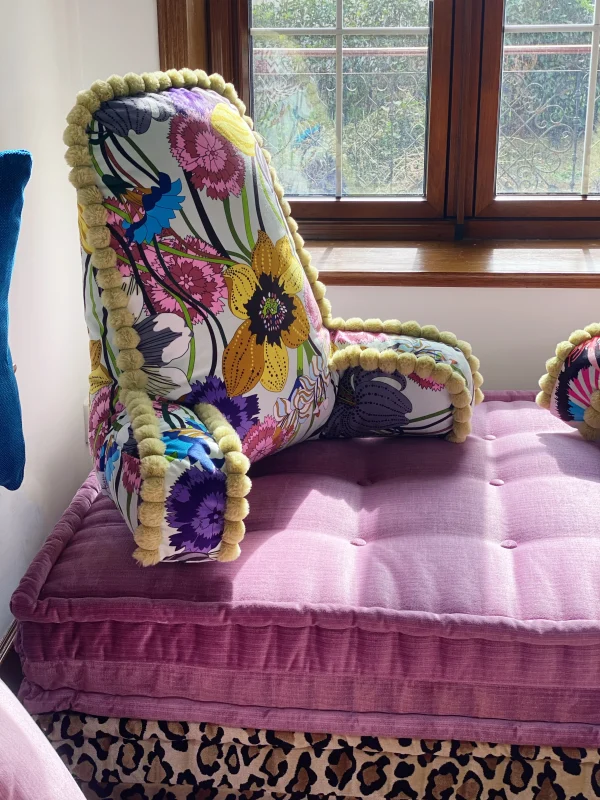 Close-up of a French Mah Jong sofa backrest with pink textured fabric and pom-pom details, set against layered cushions and vibrant throw pillows.