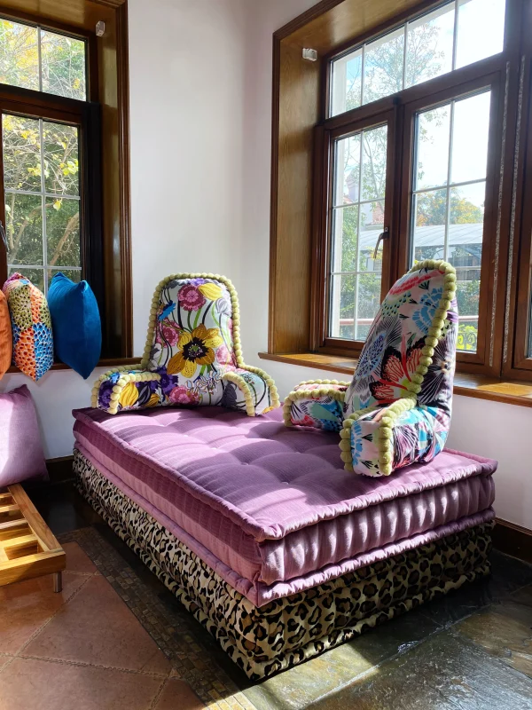 Side view of a French daybed Mah Jong sofa with a pink backrest featuring pom-pom details, paired with stacked cushions and colorful throw pillows.