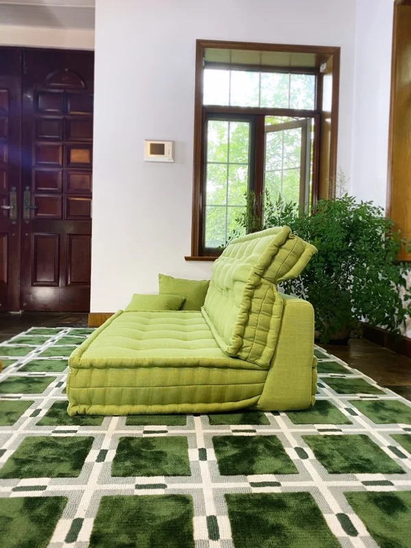 Side view of a French Mah Jong daybed in green upholstery, placed on a geometric-patterned rug with an open window in the background.