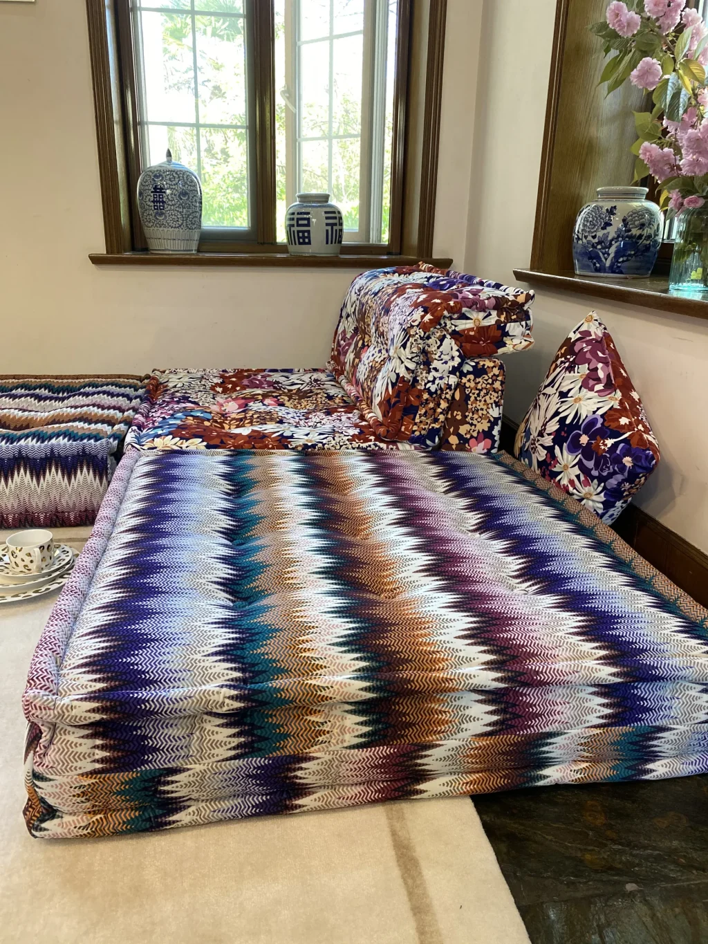 Vivid zigzag-patterned Mah Jong Sofa with floral cushions and a tea set, set against large windows.
