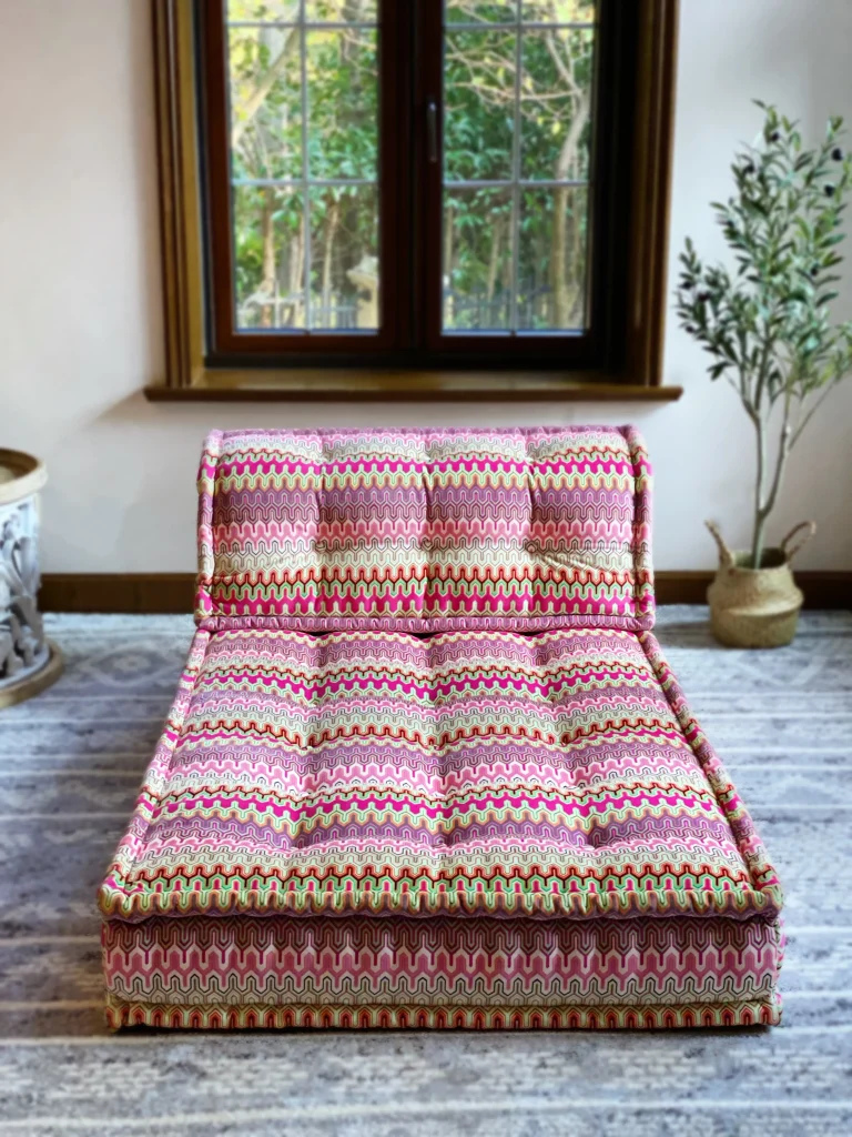 Front-facing view of the French Daybed Mah Jong sofa with bright pink and green patterned cushions, set against a window backdrop.