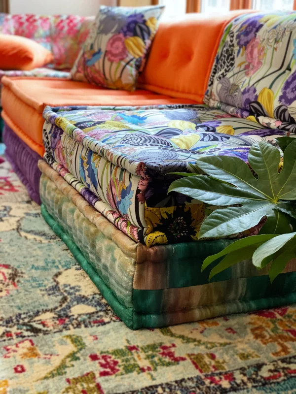 Rust-red Mah Jong daybed with a soft blue base, surrounded by neutral cushions and styled on a watercolor-inspired rug in a modern living room.