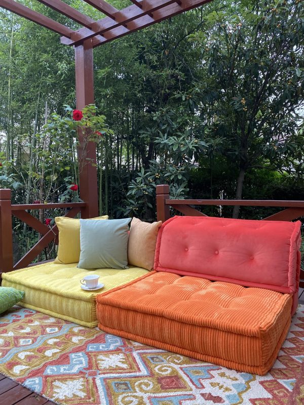 Vivid orange and yellow Mah Jong daybed placed under a wooden pergola, styled with colorful cushions and a patterned rug, surrounded by lush greenery.