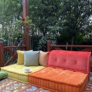 Vivid orange and yellow Mah Jong daybed placed under a wooden pergola, styled with colorful cushions and a patterned rug, surrounded by lush greenery.
