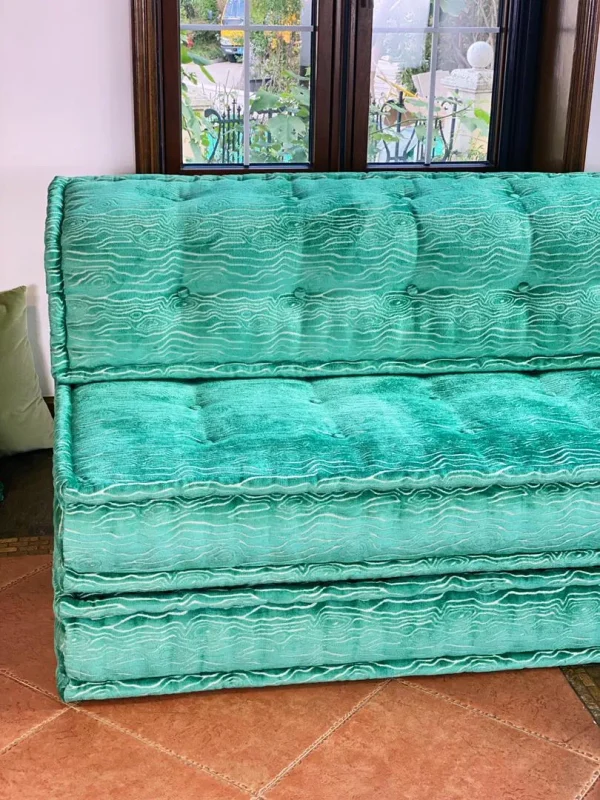 French turquoise daybed with tufted cushions styled near large windows and colorful decor accents.