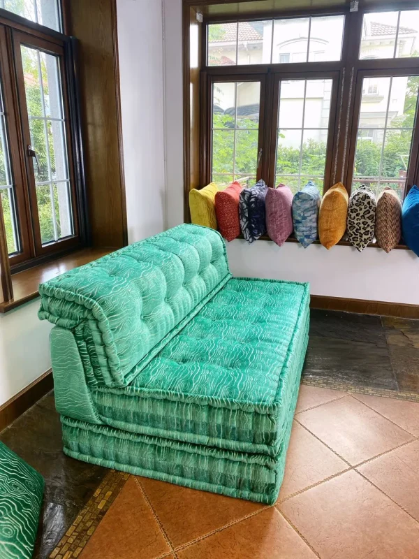 Vibrant turquoise daybed with intricate upholstery, surrounded by colorful throw pillows and natural lighting.