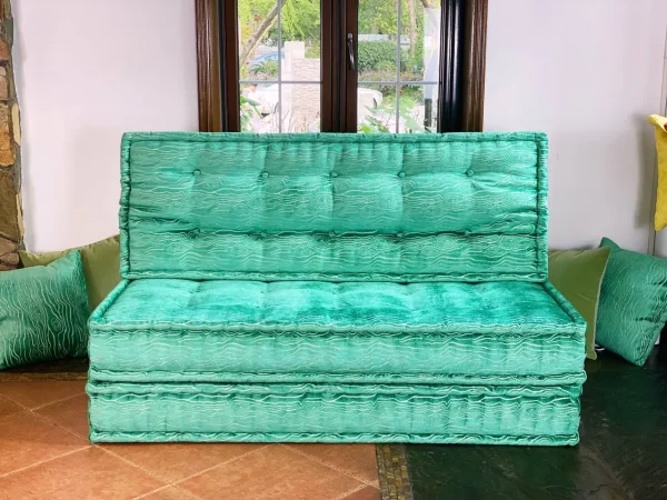 A French turquoise daybed with tufted cushions, surrounded by colorful textiles and bright natural lighting.