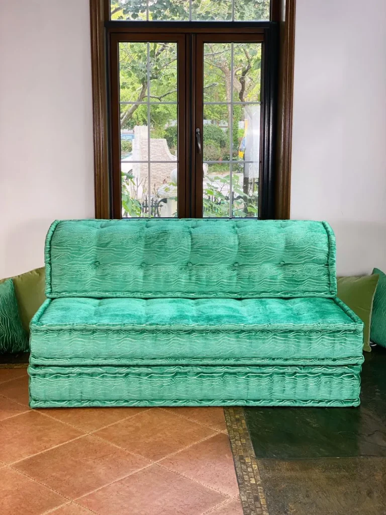 French turquoise Mah Jong daybed with a sleek and minimalist design, placed in front of a window with garden views.