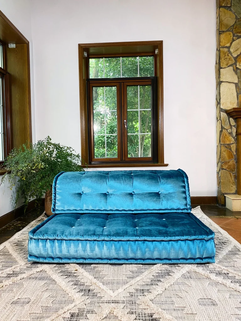 French daybed Mah Jong sofa in teal velvet upholstery, styled in a cozy living room with wooden-framed windows and greenery.