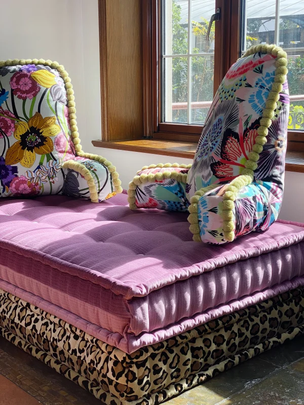 French daybed Mah Jong sofa with vibrant stacked cushions in blue, pink, and yellow patterns, with backrests and a cozy living room setup.