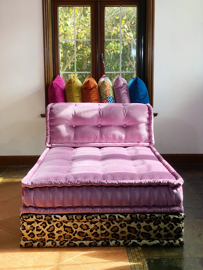 Pink tufted Mah Jong daybed with a leopard print base, framed by a sunny window and colorful pillows.