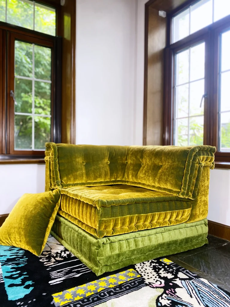 Green velvet Mah Jong daybed with a matching throw pillow on a patterned rug near a window.