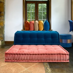 A vibrant Mah Jong-style sofa with deep blue and coral pink tufted cushions, accented with colorful throw pillows, placed by a window.