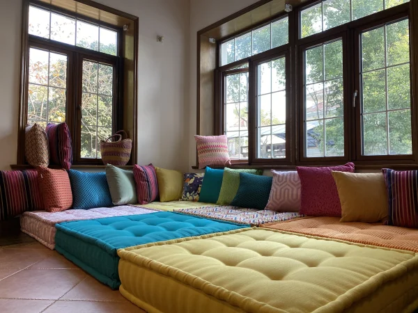 A spacious Mah Jong sofa in vibrant tones of turquoise, yellow, and orange, decorated with striped and textured pillows, beside a large window.