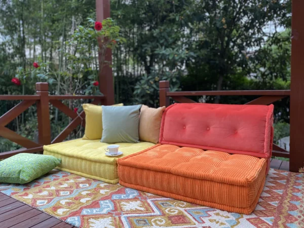 Wide view of an orange and yellow Mah Jong daybed in a garden, styled with a patterned rug and surrounded by greenery.