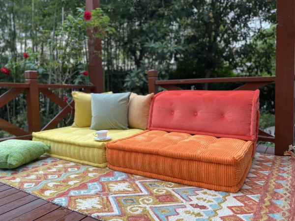 Orange and yellow Mah Jong daybed styled outdoors with floral accents, lush greenery, and a wooden pergola.