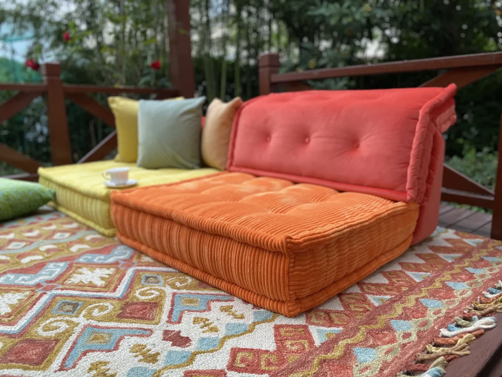 Orange and yellow Mah Jong sofa cushions placed on a colorful patterned rug, styled with a teacup and surrounded by lush greenery.