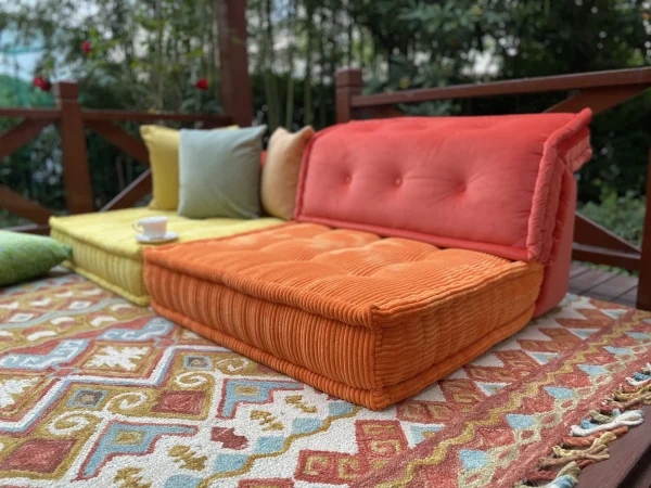 Orange and yellow Mah Jong sofa cushions placed on a colorful patterned rug, styled with a teacup and surrounded by lush greenery.