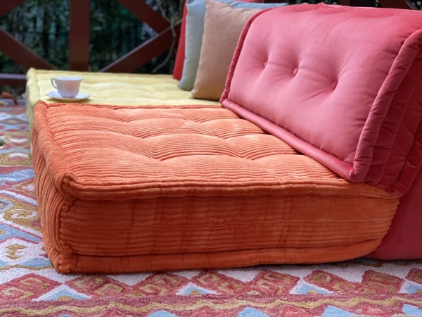 Close-up of an orange Mah Jong sofa cushion with corduroy fabric texture, placed on a colorful patterned rug outdoors.