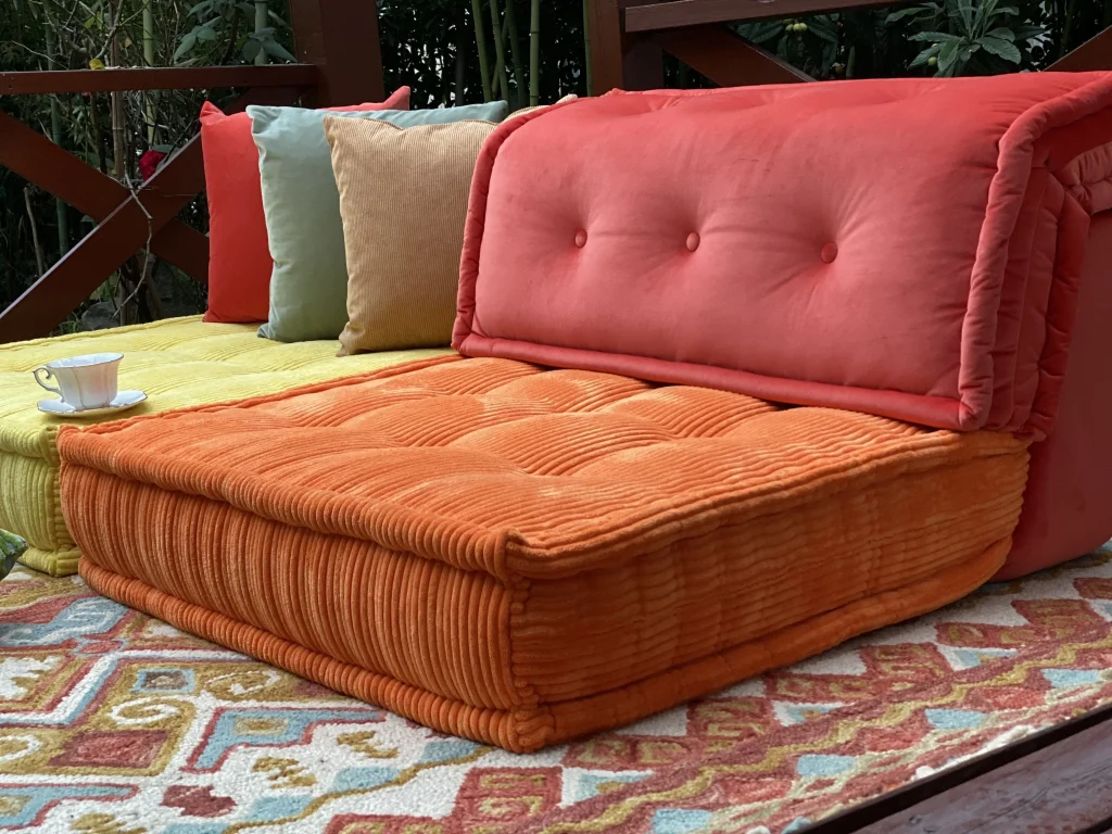 Bright orange and yellow Mah Jong sofa placed on a patterned rug under a wooden pergola, accessorized with colorful cushions and a teacup.