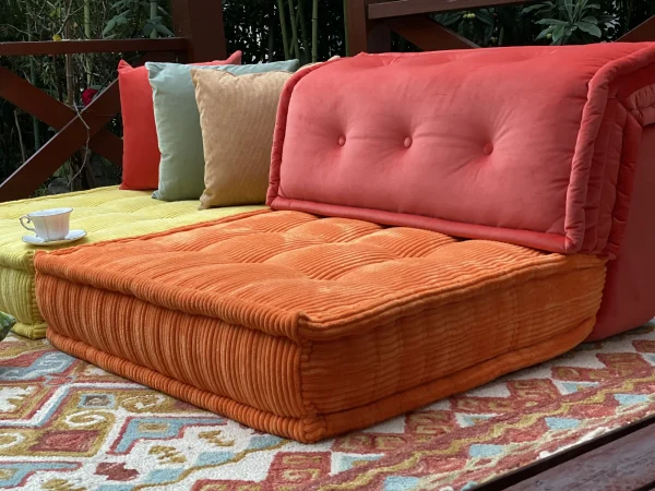 Bright orange and yellow Mah Jong sofa placed on a patterned rug under a wooden pergola, accessorized with colorful cushions and a teacup.