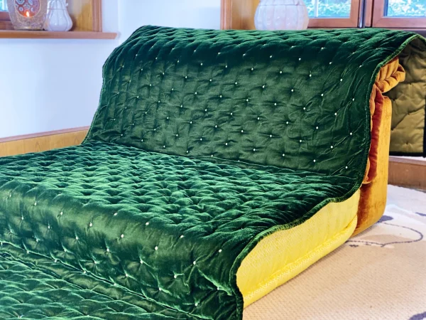 Perspective view of French daybed Mah Jong sofa, featuring a quilted green velvet cover and yellow patterned cushion.