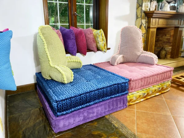 Stacked pink and yellow cushions on a French Mah Jong daybed sofa, with colorful throw pillows in the background of a modern interior.