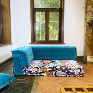 Teal velvet Mah Jong daybed with colorful floral mattress placed near wooden-framed windows.