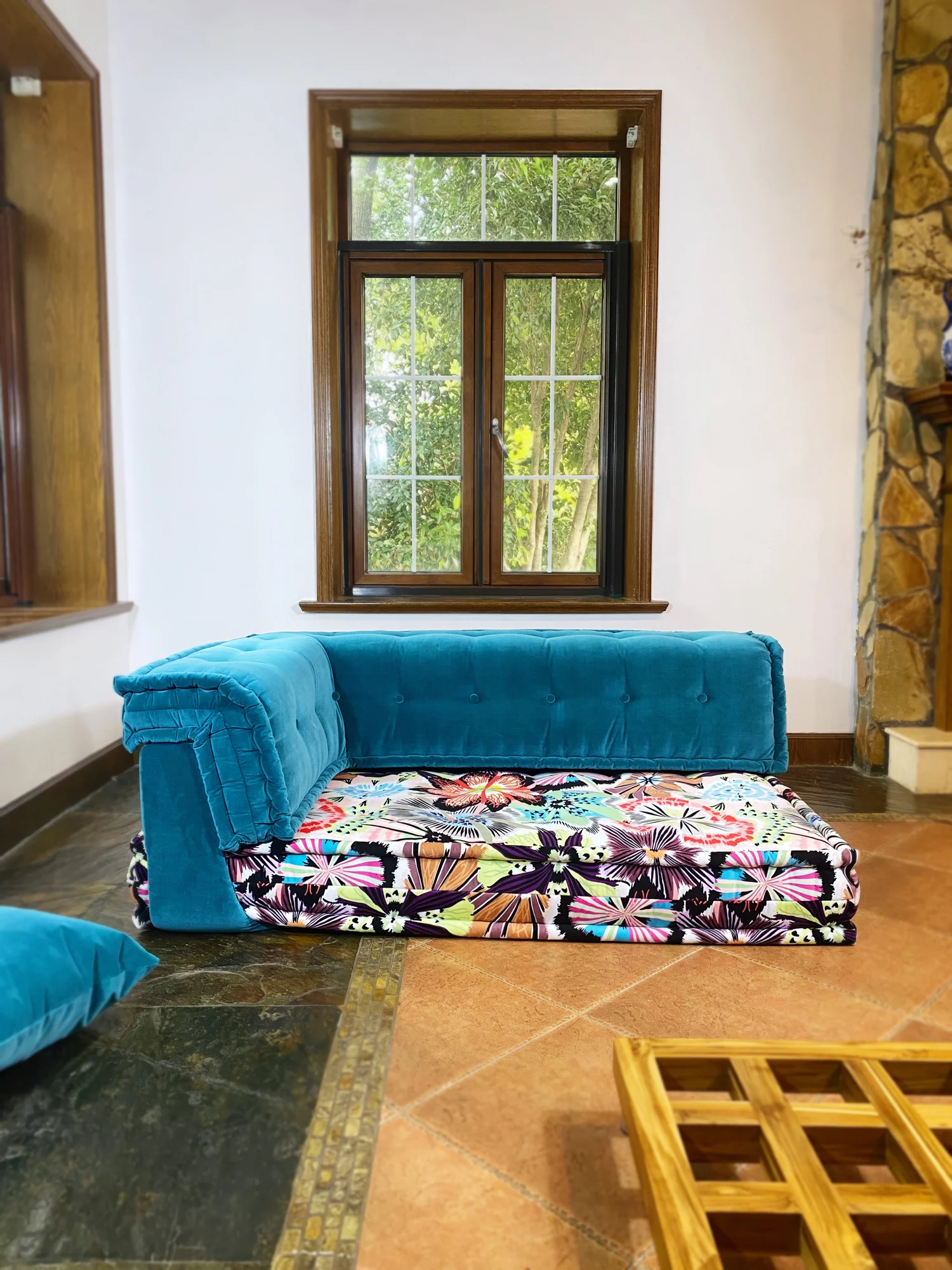 Teal velvet Mah Jong daybed with colorful floral mattress placed near wooden-framed windows.