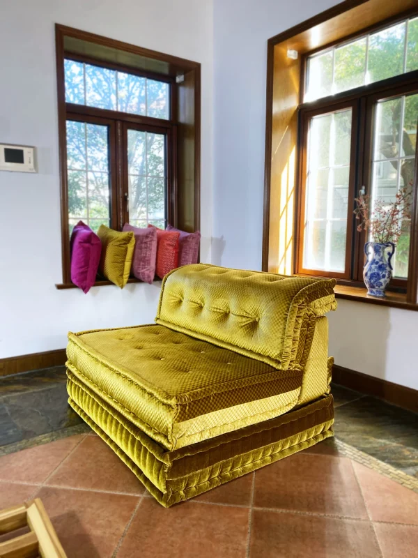 French Mah Jong daybed in golden-yellow fabric, with tufted cushions and elegant textures, styled near a window with sunlight.