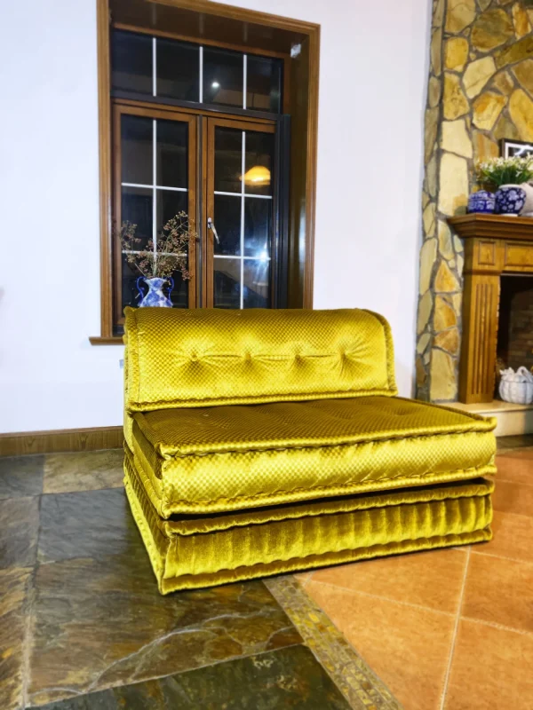 Close-up of a golden-yellow French Mah Jong daybed with rich textured fabric and a classic stone fireplace in the background.