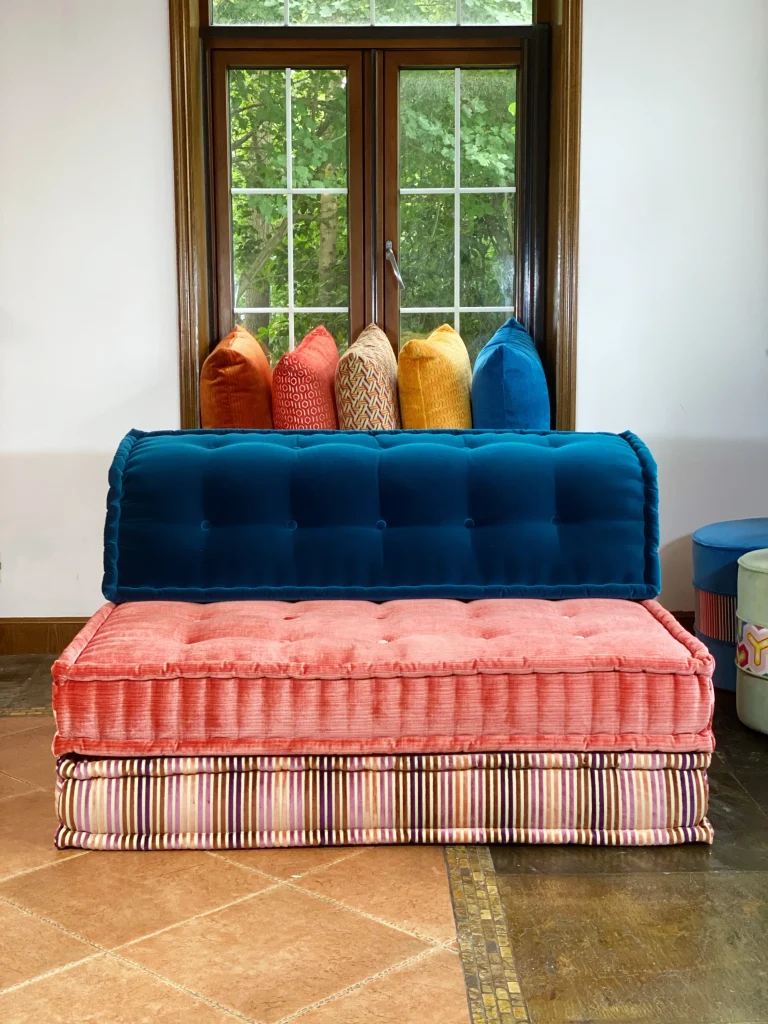 Mah Jong sofa with blue velvet backrest, pink cushions, and a striped base in a stylish room.