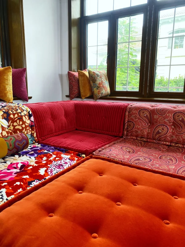 Colorful Mah Jong modular sofa setup with red paisley, floral, orange velvet, and red corduroy sections, placed in a bright living room.