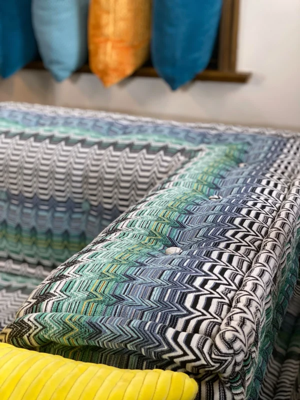 Vibrant Mah Jong sofa styled with patterned cushions, striped bolsters, and bold colors, surrounded by large windows and greenery.