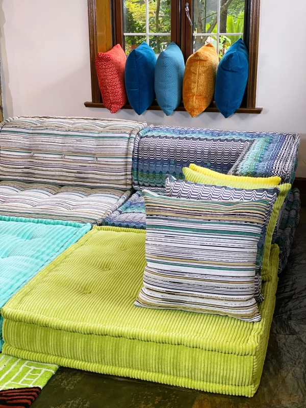 Colorful Mah Jong sofa with striped upholstery, surrounded by vibrant cushions displayed on a windowsill in a sunlit living room.