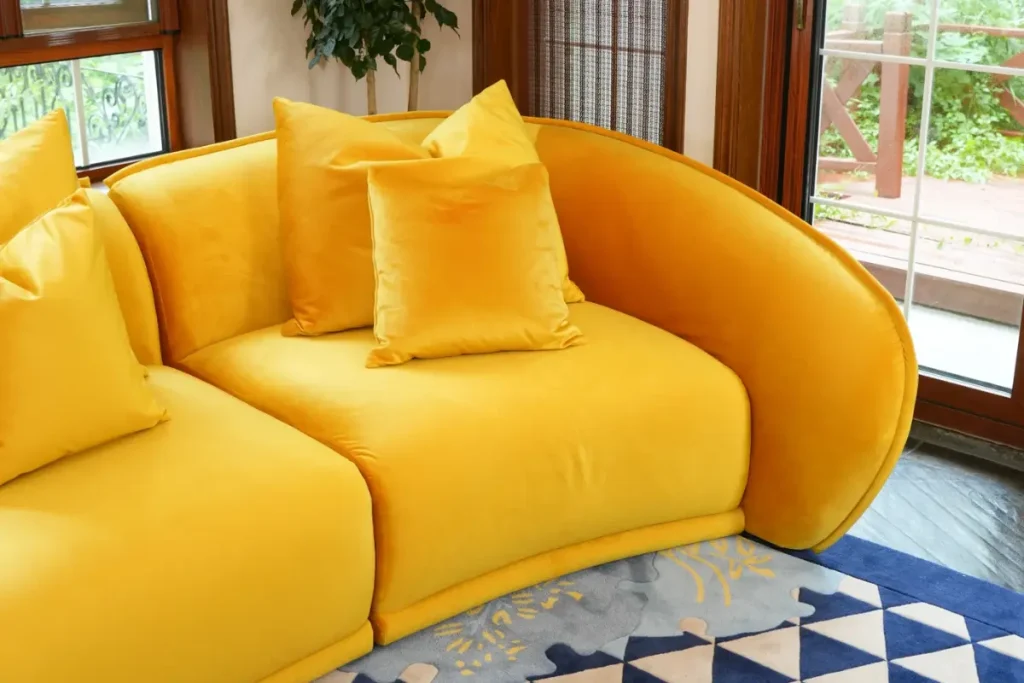 The curved arm of the Elva sofa in yellow velvet with square pillows by a window