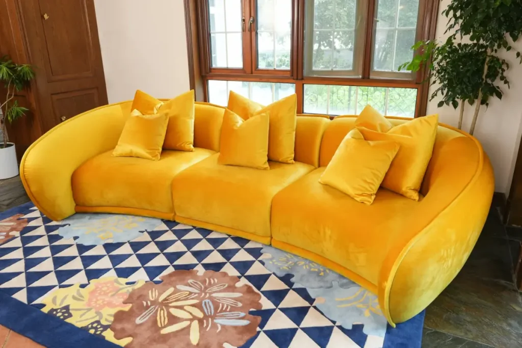 The Elva curved sofa in yellow velvet with matching pillows on a blue patterned rug