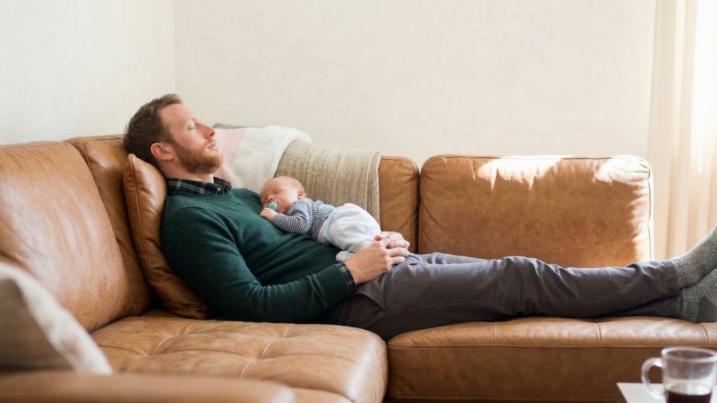 A dad and an infant sleeping on a leather couch.